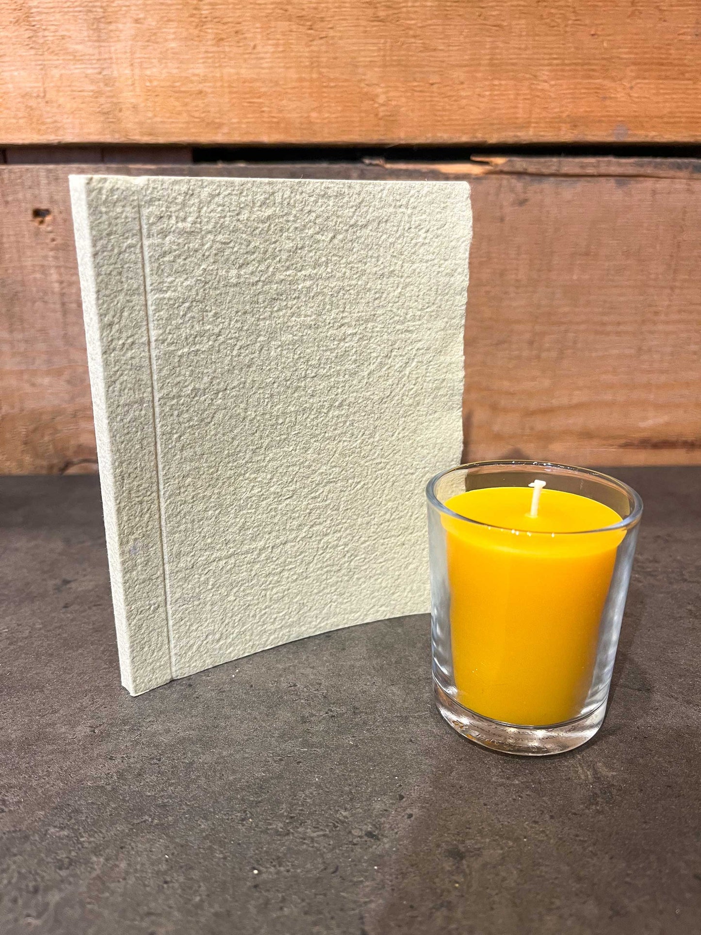 Handmade beeswax votive candle in glass cup next to pale green eco-friendly notebook