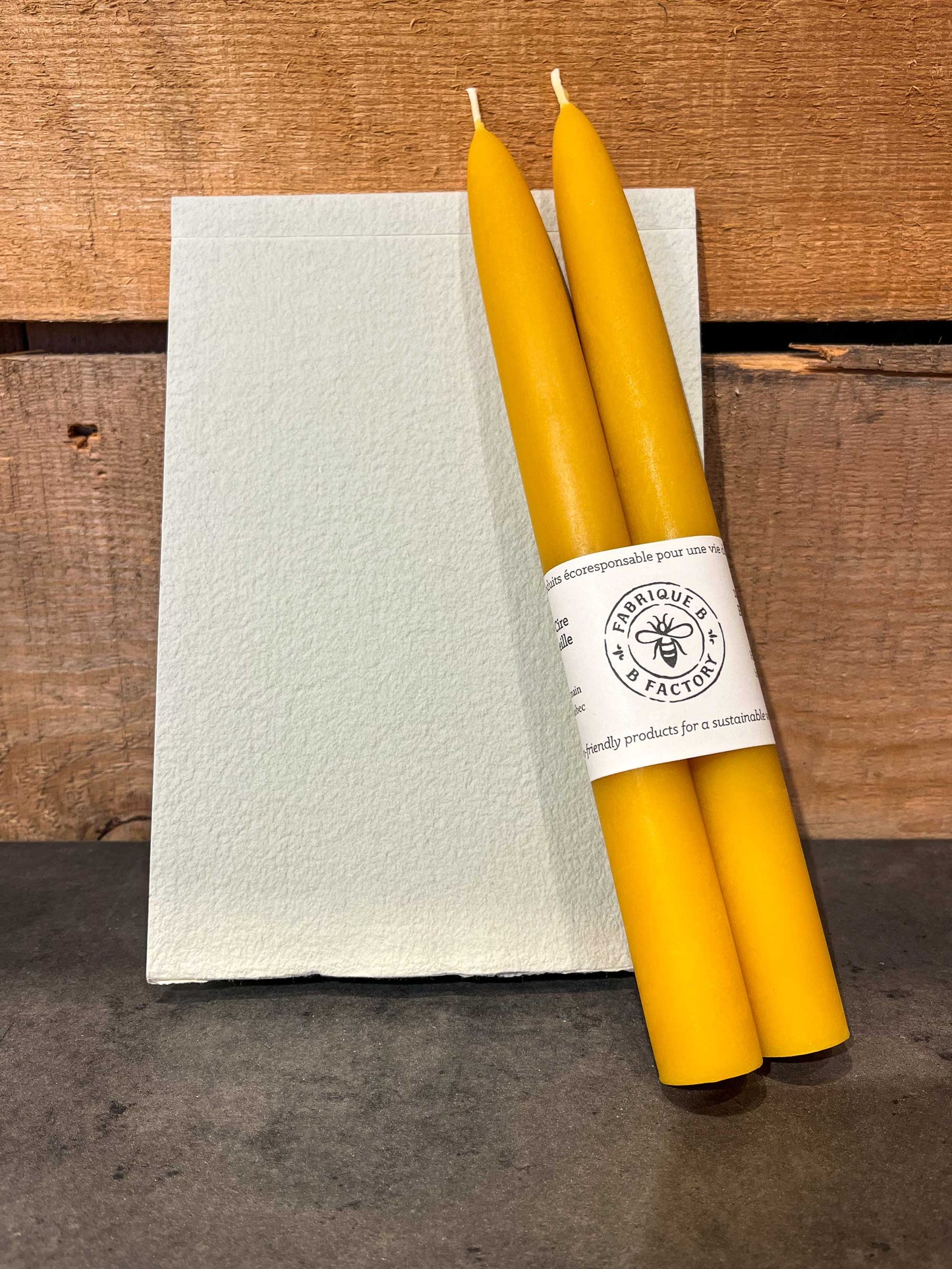 Set of 2 handmade beeswax taper candles with B Factory logo on top of beige eco-friendly notebook