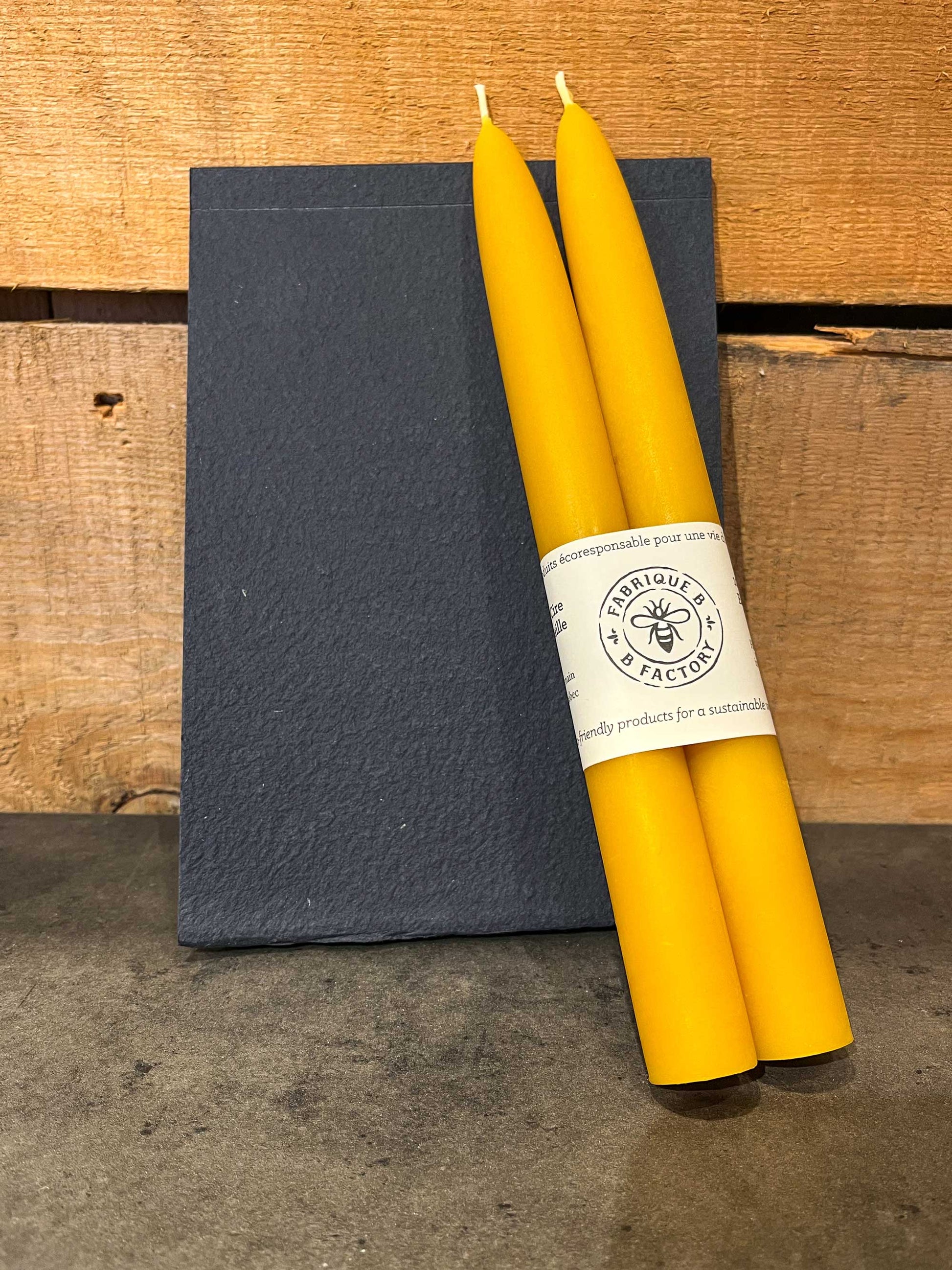 Set of 2 handmade beeswax taper candles with B Factory logo on top of blue eco-friendly notebook