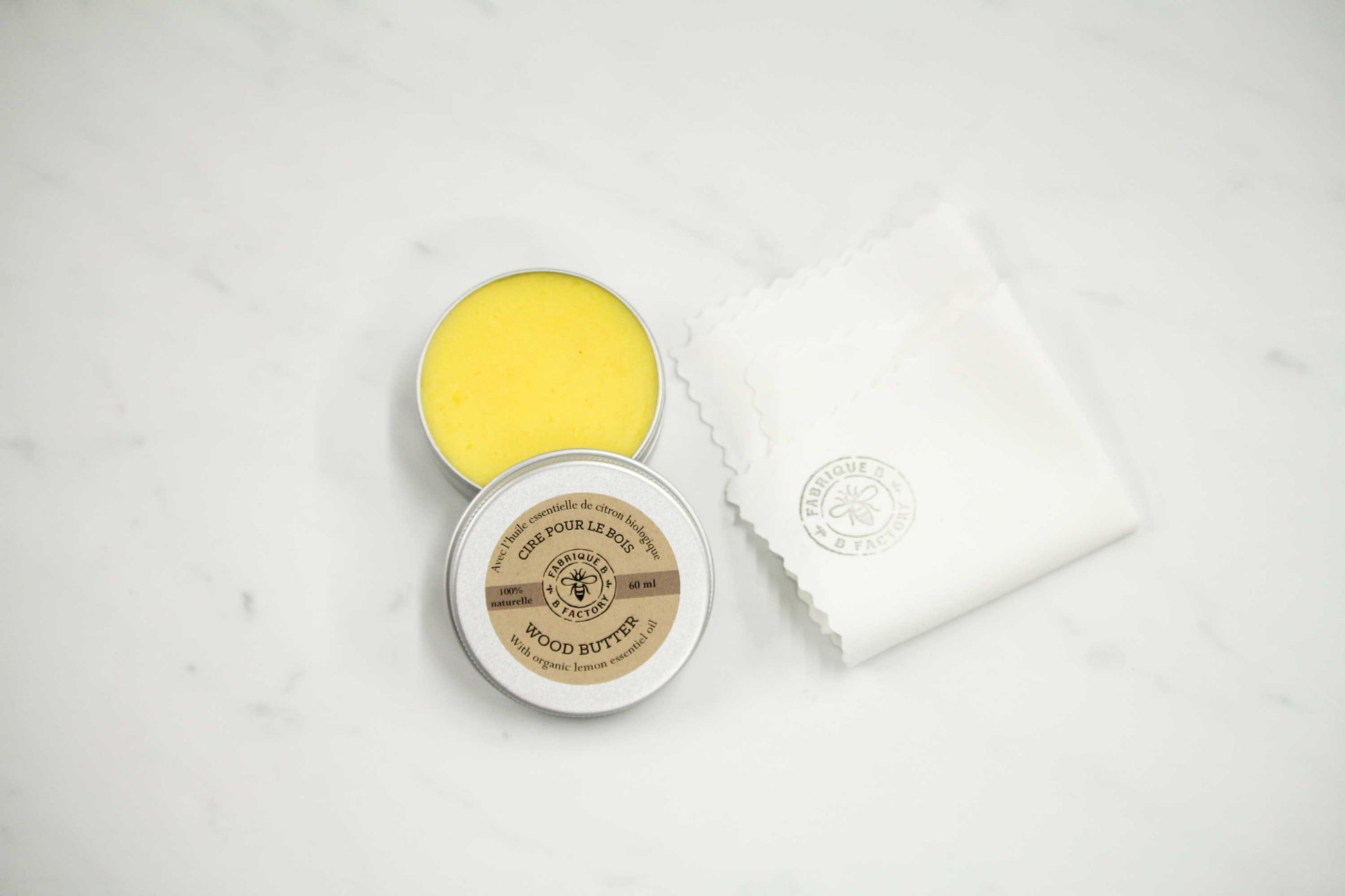 Natural beeswax wood butter in open silver tin with B Factory logo on lid next to white cloth