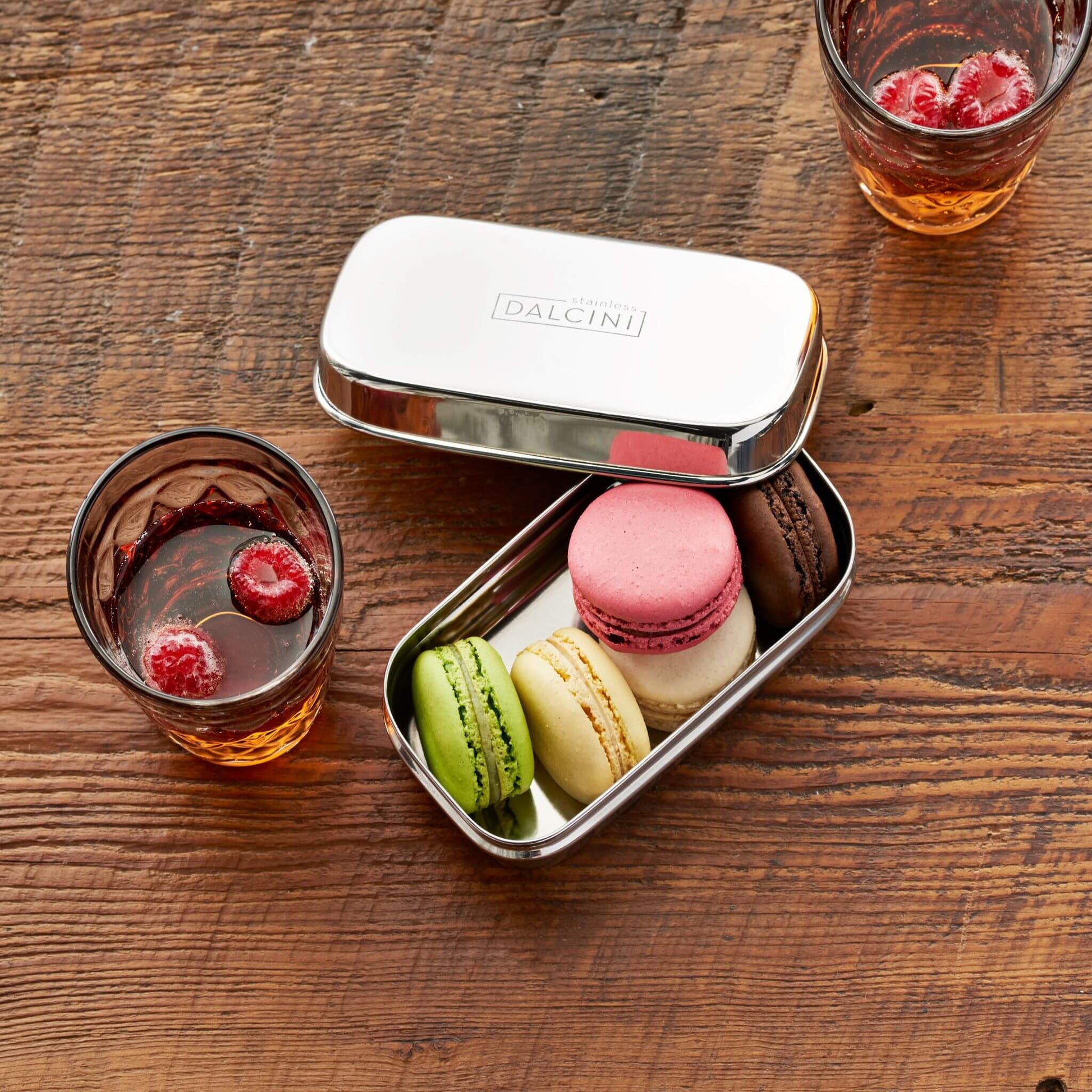 5 colourful macarons in stainless steel snack box next to glass of iced tea with raspberries