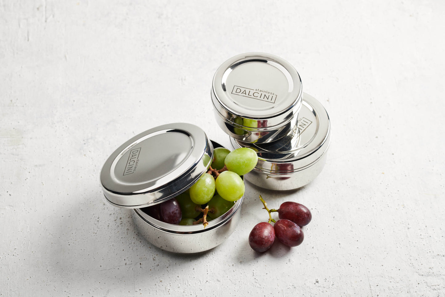 Red and green grapes coming out of round stainless steel snack box next to 2 closed stainless steel round snack boxes