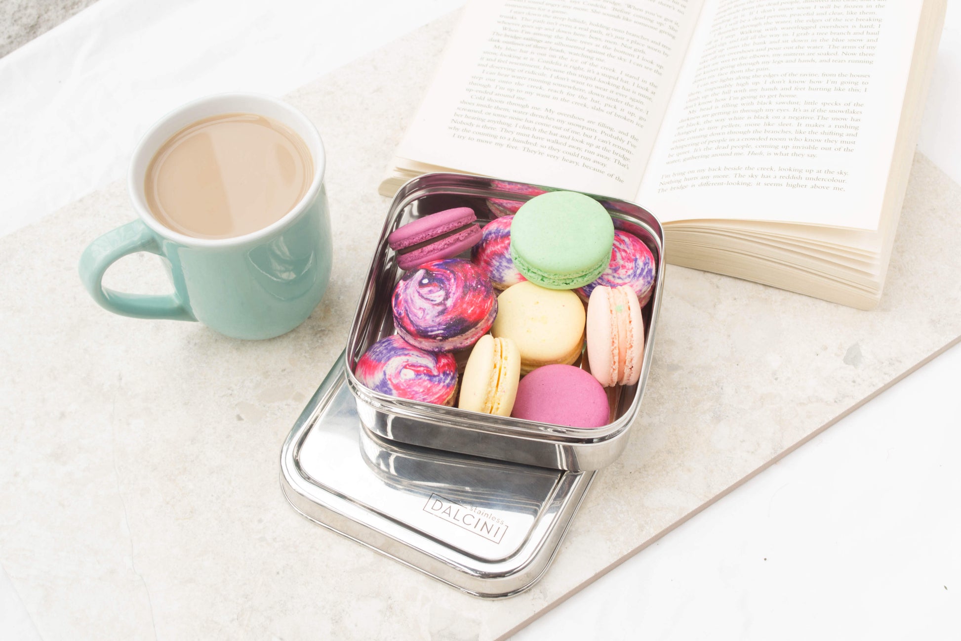 Mug of tea next to an open book and square stainless steel sandwich box filled with French macarons
