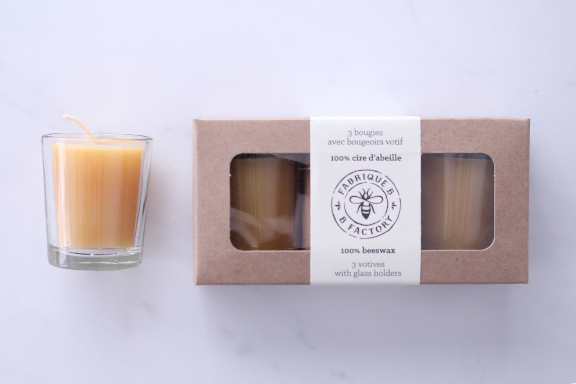Beeswax votive candle in clear glass holder next to 3 pure beeswax votive candles in gift box with B Factory logo on lid