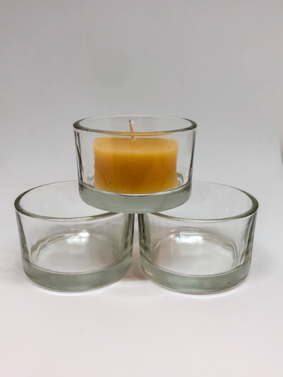 One beeswax tea light candle in clear glass cup sits on top of 2 empty clear glass tea light cups.