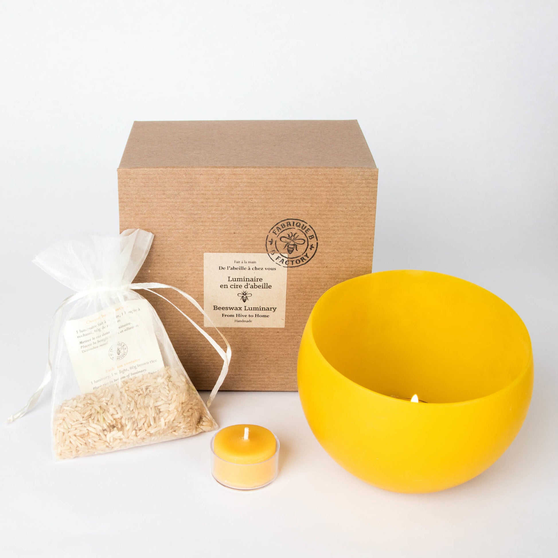 Beeswax luminary next to bag of brown rice, beeswax tea light candle in glass cup, and gift box with B Factory logo